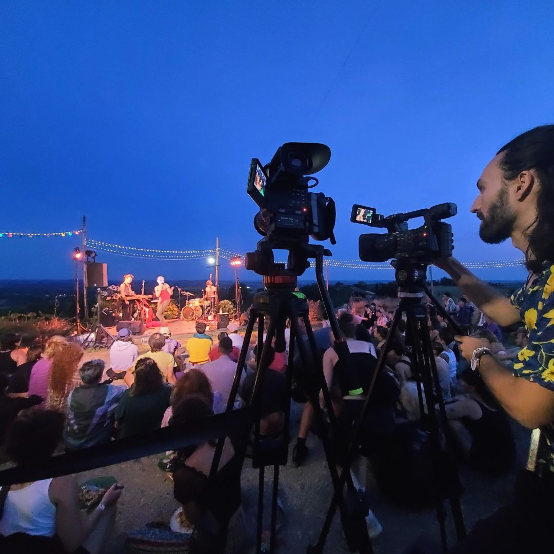 Personne en chemise bleue à fleur jaunes et cheveux longs, attentive derrière deux caméras professionnelles pointées vers une petite scène avec pour arrière plan la campagne. Sur scène, des musiciens et une drag-queen qui chante. Devant la scène, un public coloré. C'est l'heure bleue.